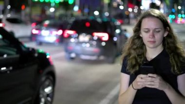 Geceleri sokakta yürürken cep telefonuyla mesajlaşan genç bir kadın.