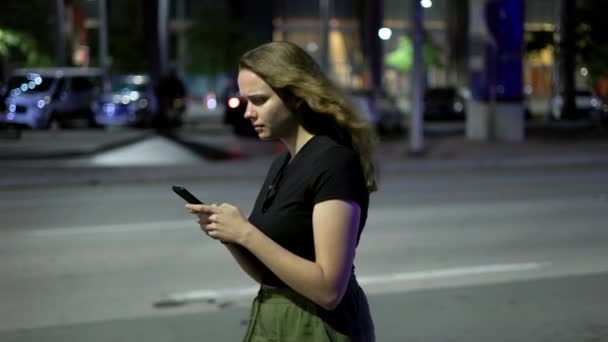 Junge Frau schreibt SMS auf ihrem Handy, während sie nachts auf der Straße spaziert — Stockvideo