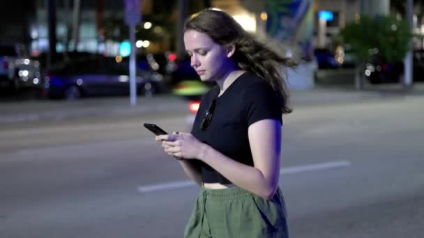Mujer joven escribiendo en su teléfono mientras camina por la calle por la noche — Vídeos de Stock
