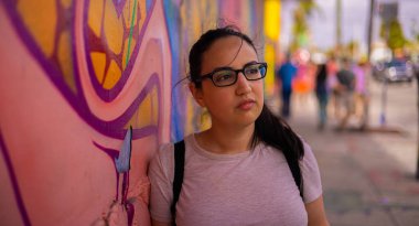 Genç ve güzel bir kadın renkli bir duvara yaslanır ve kameraya poz verir - tipik sokak tarzı.