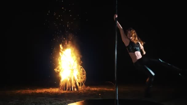 Pole dance. Halloween de sorcière. fit gymnaste féminine, en tenue en cuir noir et talons hauts, effectue des exercices acrobatiques sur mât rotatif en métal, la nuit, à la lumière d'un grand feu en arrière-plan. — Video