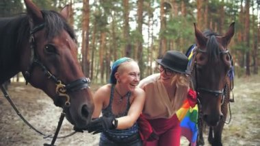 İgbt. Gökkuşağı bayrağı. Aynı cinsiyette aşk. Genç lezbiyen eşcinsel çift ortak hobileri olan ata biniyorlar. Kadınlar ormanda atlarının yanında sarılıyor, eğleniyorlar.