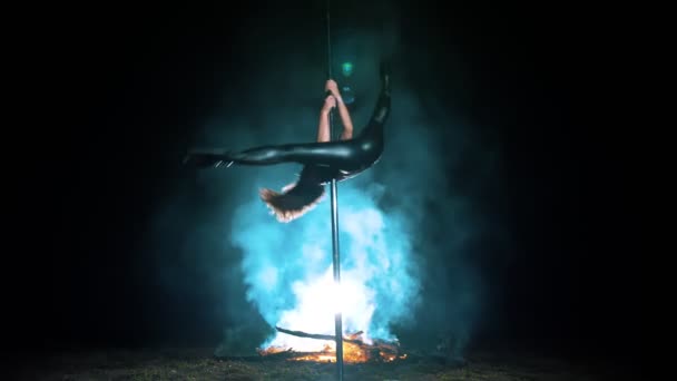 ポールダンスだ。魔女ハロウィーン。黒革の装身具とハイヒールで女性の体操選手にフィットし金属回転極でアクロバティックな練習を夜に大火災を背景にして行います. — ストック動画