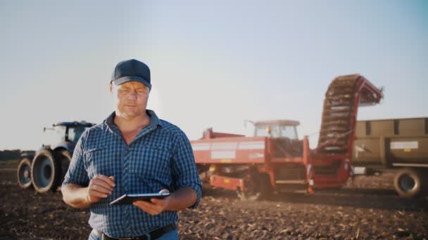 Збирання картоплі. фермер використовує цифровий планшет. на фермерському полі, на фоні сільськогосподарської техніки. трактор з картопляним комбайном. розумне сільське господарство. технології вирощування — стокове відео