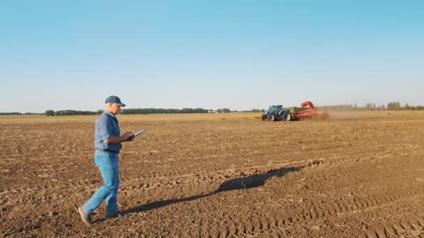 Cosecha de patatas. agricultor, sosteniendo una tableta digital en sus manos, camina a través de un campo recién arado contra el telón de fondo de la maquinaria agrícola de trabajo, cosechadoras de patatas — Vídeo de stock