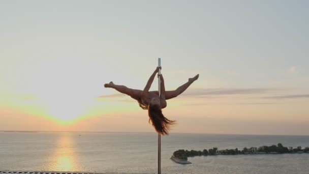 Baile pole. silueta. gimnasta femenina en forma, en maillot beige, realiza ejercicios acrobáticos en poste giratorio de metal, en techo de rascacielos, con fondo de vista mar amanecer — Vídeos de Stock