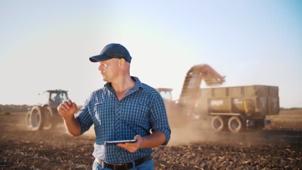 Sklizeň brambor. zemědělec používající digitální tablet. na farmářském poli, na pozadí zemědělských strojů. Traktor se sklízečem brambor. chytré zemědělství. zemědělské technologie — Stock video