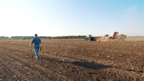 Raccolta delle patate. contadino, tenendo in mano una tavoletta digitale, cammina attraverso un campo appena arato sullo sfondo della lavorazione di macchine agricole, raccoglitori di patate — Video Stock