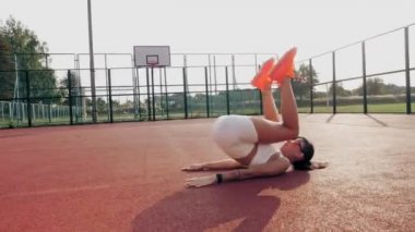 Spor salonu açık havada. Beyaz bluzlu, şortlu, turuncu spor ayakkabılı ve güneş gözlüklü sportif bir kadın, turuncu sahada beyaz işaretli çeşitli egzersizler yapıyor. Yaz günü. Açık hava sporları