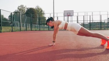 Spor salonu açık havada. Beyaz bluzlu, şortlu, turuncu spor ayakkabılı ve güneş gözlüklü sportif bir kadın, turuncu sahada beyaz işaretli çeşitli egzersizler yapıyor. Yaz günü. Açık hava sporları