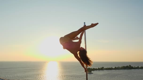 Paaldansen. silhouet. fit vrouwelijke gymnaste, in beige maillot, voert acrobatische oefeningen op metalen roterende paal, op wolkenkrabber dak, met zee zonsopgang uitzicht achtergrond — Stockvideo