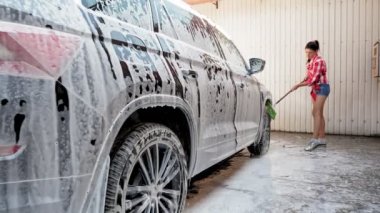 Araba yıkama. Şortlu ve gömlekli genç bir kadın, sabunlu köpükle kaplı bir arabayı yıkıyor, özel bir fırçayla, oto yıkamada, dışarıda. Araba Temizleme