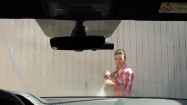 Bir kadın arabanın ön camını temiz suyla yıkıyor yüksek basınçlı spreyle, dışarıda, oto yıkamada. Araba temizliği. Otomobilin içinden görüntüle — Stok video