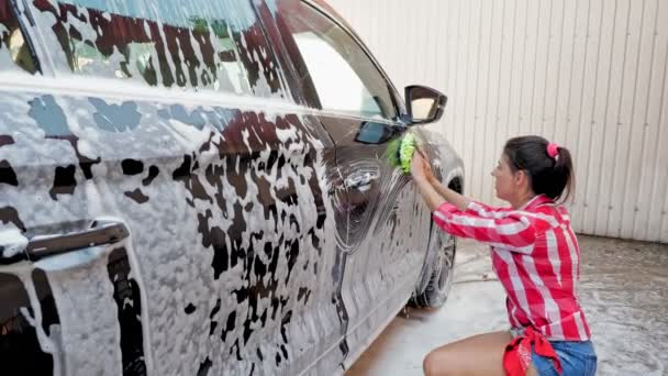 Lavado de coches. mujer joven, en pantalones cortos y camisa, está lavando un coche, cubierto con espuma jabonosa, con un paño de baño, en auto servicio de lavado de coches, al aire libre. Limpieza de coches — Vídeos de Stock