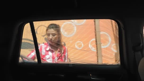 Lavado de coches. mujer joven está lavando un coche, cubierto con espuma jabonosa, con un paño, en auto servicio de lavado de coches, al aire libre. Limpieza de coches. Vista desde el interior del automóvil — Vídeos de Stock