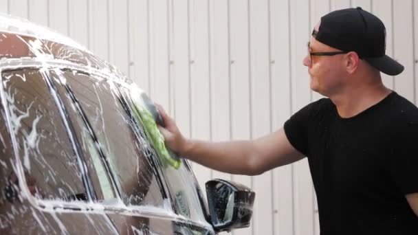 Autolavaggio. Un uomo, in maglietta nera, è lavaggio auto, coperto con schiuma saponata, con un panno, a self-service car wash, all'aperto. Pulizia auto — Video Stock