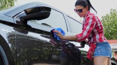 Araba yıkama, araba silme. Şortlu ve gömlekli genç bir kadın, dışarıda bir bez parçasıyla otomobili siliyor, yıkandıktan sonra, oto yıkamada. Araba Temizleme.