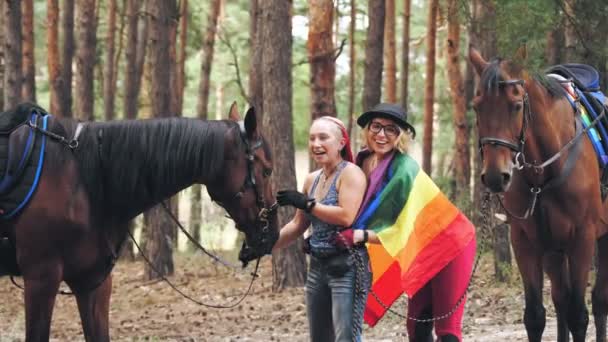 Lgbt. bandera del arco iris. amor del mismo sexo. joven pareja lesbiana del mismo sexo se dedica a afición conjunta, equitación. mujeres abrazándose, divirtiéndose, junto a sus caballos, en el bosque — Vídeo de stock