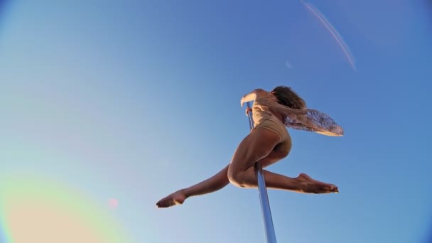Direk dansı. Bej mayo giyen formalı kadın jimnastikçi, mavi gökyüzüne karşı metal dönen direkte akrobatik egzersizler yapıyor. aşağıdan yukarıya görünüm — Stok video