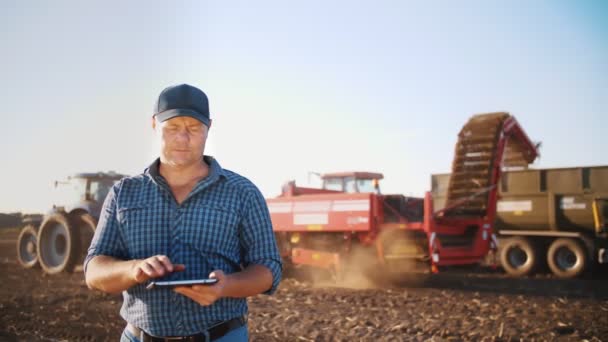 Raccolta delle patate. agricoltore utilizzando tablet digitale. sul campo agricolo, sullo sfondo macchine agricole. trattore con raccoglitore di patate. agricoltura intelligente. tecnologie agricole — Video Stock