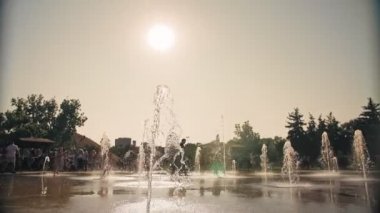 Çeşme. Fıskiye fıskiyeleri. Arka planda bulanık çocuklar, su akıntıları arasında eğleniyorlar, şehir parkında yüksek çeşme jetleri, sıcak yaz günü.