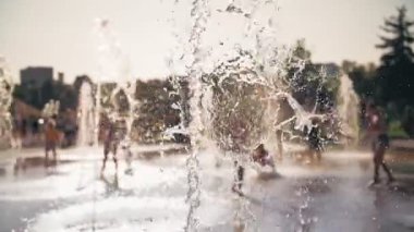 Çeşme. Yakın plan. Fıskiye fıskiyeleri. Arka planda bulanık çocuklar, su akıntıları arasında eğleniyorlar, şehir parkında yüksek çeşme jetleri, sıcak yaz günü.