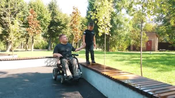 Vaderdag. persoon met een handicap. Papa en zijn schattige zoontje lopen in het park, op zomerdag. Papa is een rolstoelgebruiker. mensen met bijzondere behoeften. — Stockvideo