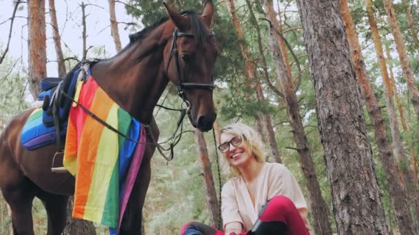 Regnbågsflagga. Ridning. vacker ryttare flicka, med lgbt regnbåge flagga, poserar bredvid sin häst, medan du sitter och vilar under promenader i skogen. — Stockvideo