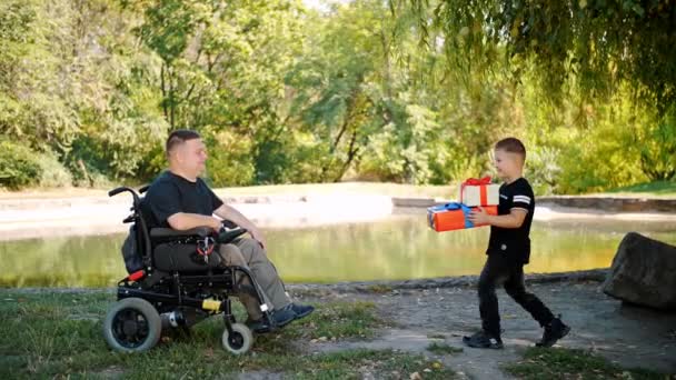 Día de los padres. pequeño niño lindo le da regalos a su padre. un hombre es una persona con discapacidad. usuario de silla de ruedas. personas con necesidades especiales. — Vídeos de Stock