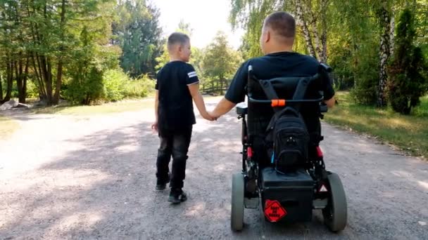 Dzień ojców. osoba niepełnosprawna. Tata i jego słodki synek spacerują po parku, w letni dzień. Tata używa wózków inwalidzkich. osoby o szczególnych potrzebach. — Wideo stockowe