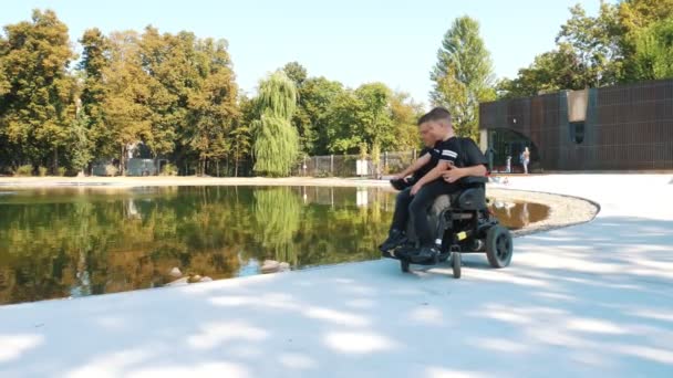 Día de los padres. persona con una discapacidad. Papá y su pequeño hijo lindo están caminando en el parque, en el día de verano. Papá es un usuario de silla de ruedas. personas con necesidades especiales. — Vídeo de stock