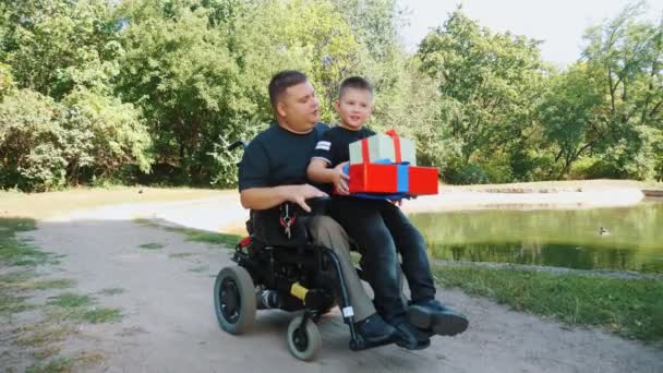 Día de los padres. pequeño niño lindo se sienta en su regazo padres con regalos en sus manos. un hombre es una persona con discapacidad. usuario de silla de ruedas. personas con necesidades especiales. — Vídeos de Stock