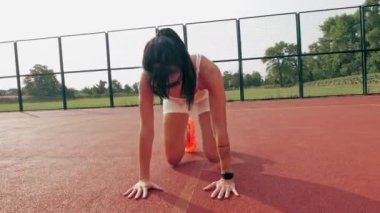 Spor salonu açık havada. Beyaz bluzlu, şortlu, turuncu spor ayakkabılı ve güneş gözlüklü sportif bir kadın, turuncu sahada beyaz işaretli çeşitli egzersizler yapıyor. Yaz günü. Açık hava sporları