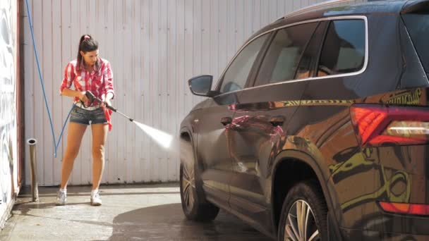 Egy csinos nő lemossa az autót tiszta vízzel, nagynyomású sprayvel, odakint, az önkiszolgáló autómosóban. Autótisztítás. — Stock videók