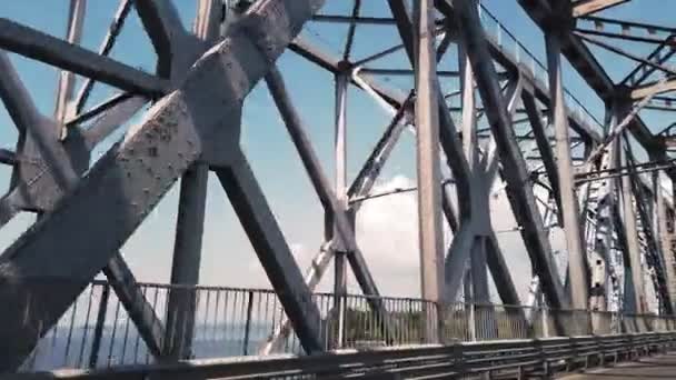 Ponte di metallo. costruzioni in ferro grezzo contro il cielo blu e il fiume, girato in movimento da una macchina. vista laterale. Ponte ferroviario e stradale attraverso il fiume. Silhouette di traversata travi in acciaio. — Video Stock