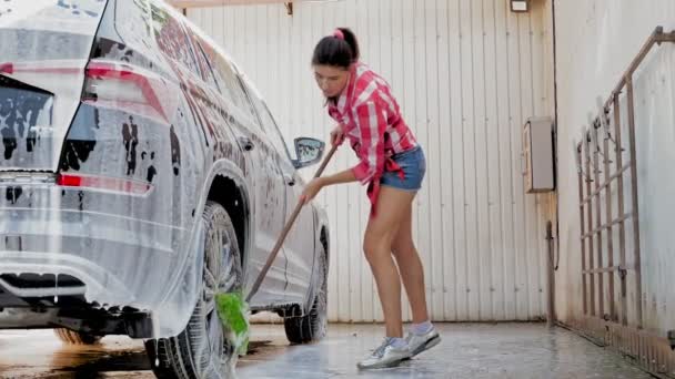 Araba yıkama. Şortlu ve gömlekli genç bir kadın, sabunlu köpükle kaplı bir arabayı yıkıyor, özel bir fırçayla, oto yıkamada, dışarıda. Araba Temizleme — Stok video