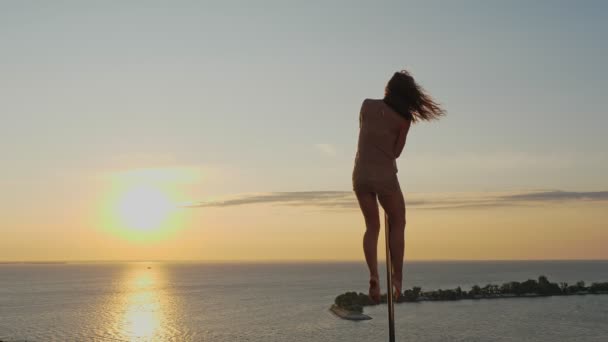 Poldansen. silhuett. passar kvinnliga gymnast, i beige leotard, utför akrobatiska övningar på metall roterande stolpe, på skyskrapa tak, med havsutsikt soluppgång bakgrund — Stockvideo