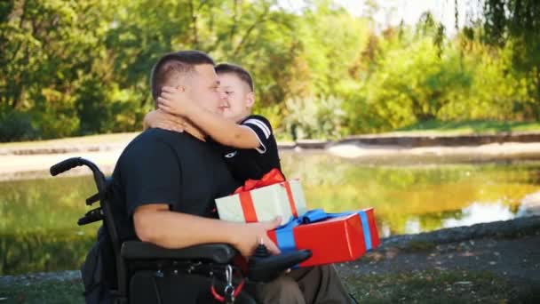 Dzień ojców. Mały, słodki chłopiec daje prezenty swojemu tacie. mężczyzna jest osobą niepełnosprawną. użytkownik wózka inwalidzkiego. osoby o szczególnych potrzebach. — Wideo stockowe