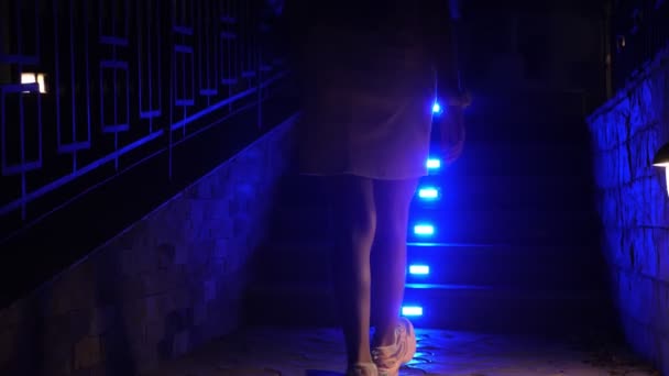 Upplysta trappsteg. närbild. silhuett av kvinnliga ben, i sneakers, klättring trappan, med klarblå belysning, på natten, i trädgården av hotellkomplexet. sommarnatt. — Stockvideo