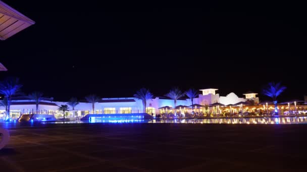Gece tropikal otelin manzarası. Zaman turları. Akşam, gece manzarası. Altın ve maviyle aydınlatılmış palmiye ağaçları olan bir otel kompleksi. İnsanlar bilardo masalarında oturuyorlar. — Stok video
