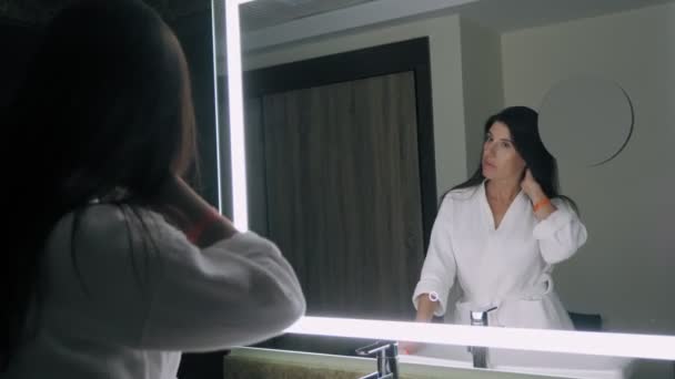 Жінка перед дзеркалом. красива молода жінка в білому халаті, стоїть перед дзеркалом і оглядає, захоплюється її відображенням — стокове відео