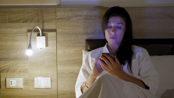 Πριν κοιμηθώ. Μια νεαρή γυναίκα με λευκό μπουρνούζι ξαπλώνει στο κρεβάτι και χρησιμοποιεί smartphone. Δίπλα στο κρεβάτι, είναι αναμμένο το φωτάκι ή το κομοδίνο. αργά το βράδυ ή τη νύχτα — Αρχείο Βίντεο