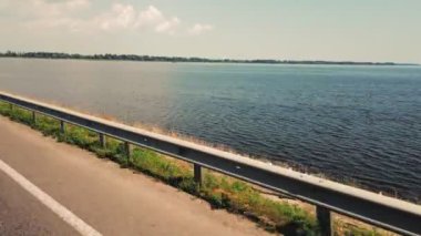 Yolculuk. Sürücü arabasının manzarası. Araba nehir ya da göl boyunca hız yapıyor. yan pencere hareketi görünümü. Nehrin karşısında otoyolu olan bir baraj. Araba yolda gidiyor, barajdan geçiyor. Hareketli manzara.