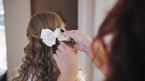 Comb child hair. close-up. mom is doing hair to her little daughter, attaches a bow to her hair. woman and little girl stand in front of the mirror. back view — Stockvideo