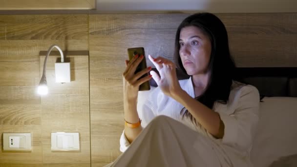 Перед сном. молода жінка в білому халаті лежить в ліжку і використовує смартфон. поруч з ліжком ввімкнено нічне світло або приліжкові бра. пізній вечір або ніч — стокове відео