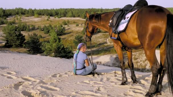 Ιππασία. Αγάπης αλόγων. νεαρή γυναίκα και το άλογό της, αναπαύονται σε αμμώδη λόφο, πανύψηλα πάνω από πευκοδάσος, το ηλιοβασίλεμα, σε ζεστές ακτίνες του ήλιου το καλοκαίρι. φιλία μεταξύ ανθρώπου και αλόγου. — Αρχείο Βίντεο