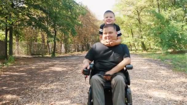 Día de los padres. persona con una discapacidad. Papá y su pequeño hijo lindo están caminando en el parque, en el día de verano. Papá es un usuario de silla de ruedas. personas con necesidades especiales. — Vídeos de Stock