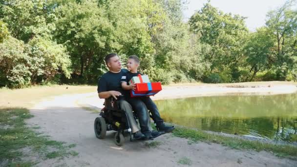 Dzień ojców. mały uroczy chłopiec siedzi na kolanach swoich ojców z prezentami w rękach. mężczyzna jest osobą niepełnosprawną. użytkownik wózka inwalidzkiego. osoby o szczególnych potrzebach. — Wideo stockowe