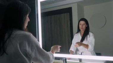 woman in front of mirror. beautiful young woman in a white bathrobe, turns off the light on mirror, stands in front of it and examines, admires her reflection.