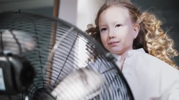 Szőrszálhasogatás a szélben. közelkép, aranyos kislány szórakozik.Ott áll a ventilátor előtt, a levegő áramlik a göndör haja, mintha a szél — Stock videók